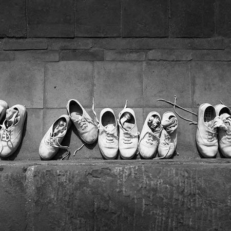 Auf einem Weg an einer Mauer stehen viele weiße Schuhpaare, das Bild ist schwarz weiß