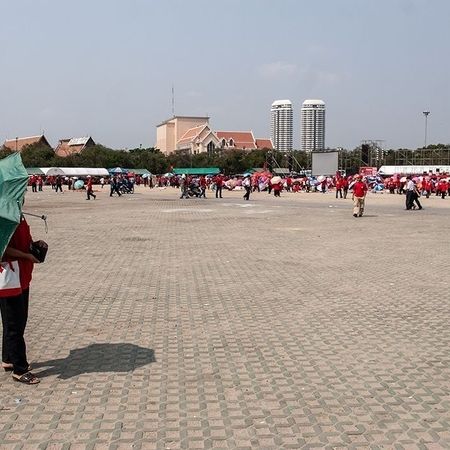 Auf einem großen Platz sind viele Personen mit roten Oberteilen verteilt, einer hat einen grünen Regenschirm über die Schulter gelehnt