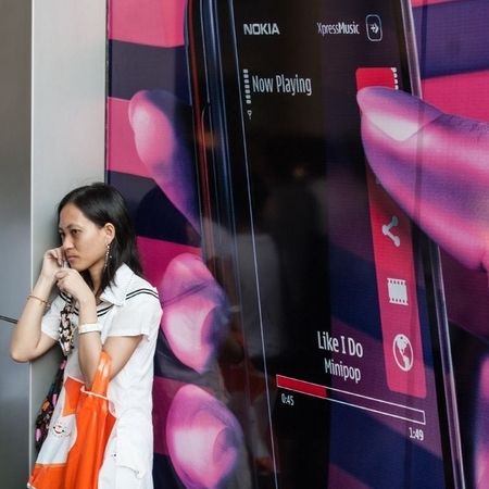 An ein Plakat mit einem Handy auf pink / violettem Hintergrund steht eine telefonierende Frau gelehnt