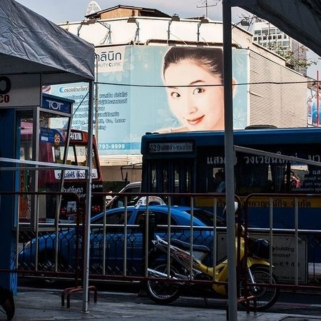 Hinter einer metallenen Absperrung ist eine dicht befahrene Straße, hinter welcher ein Gebäude mit Werbebild einer lächelnden Frau zu sehen ist