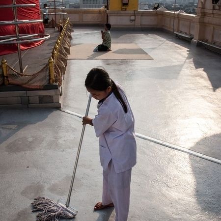 Eine Frau putzt ein mit Fahnen und Statuen versehenes Flachdach, ein Junge kniet auf einem Teppich und betet