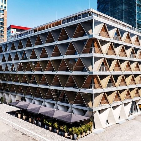 Drohnenaufnahme von einem modernen Gebäude mit geometrischer Fassade und Glasfenstern vor klarem blauen Himmel
